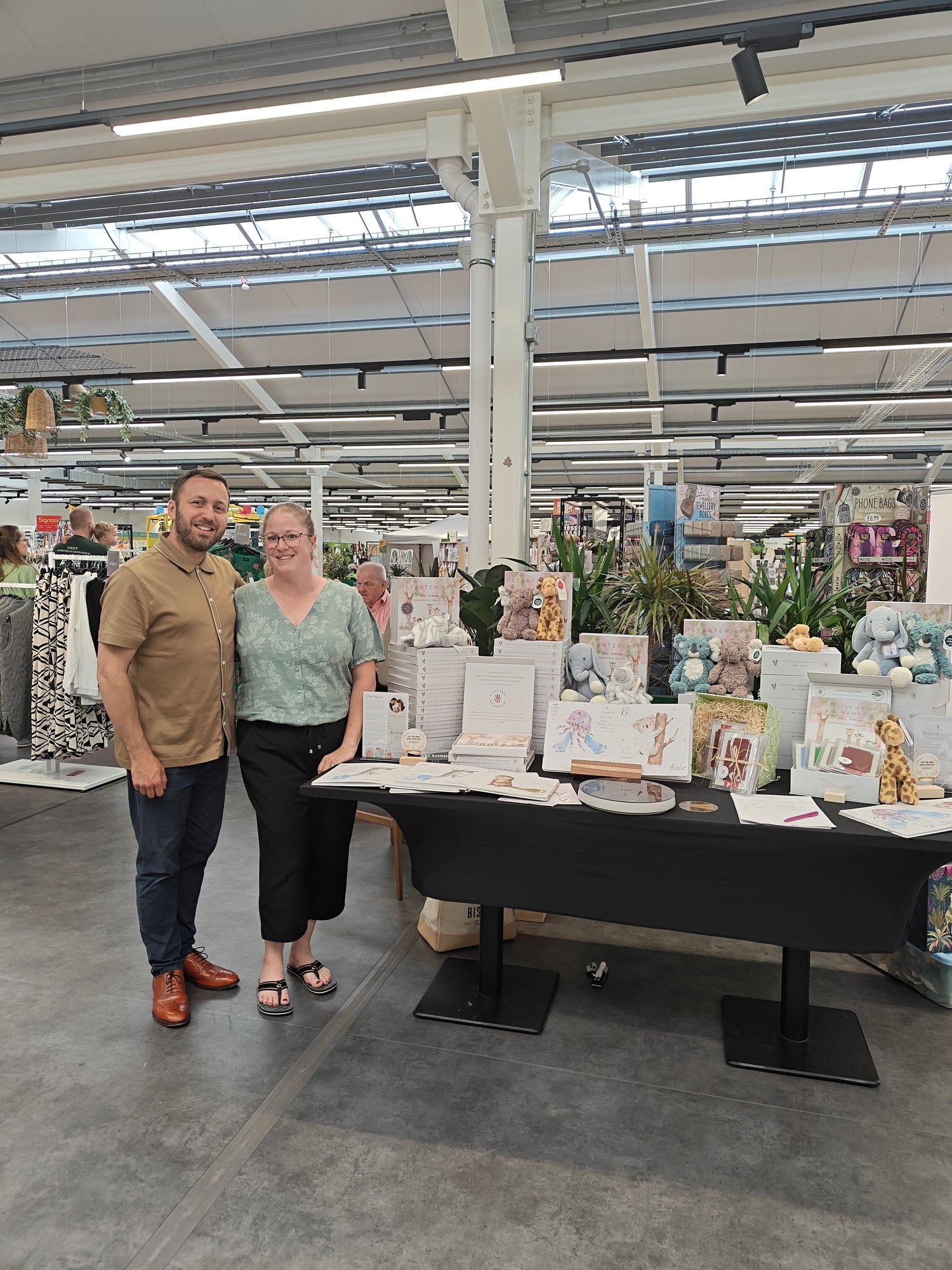 Two people standing in a store with a display of products on tables. Small business pop up in Dobbies Garden Centre